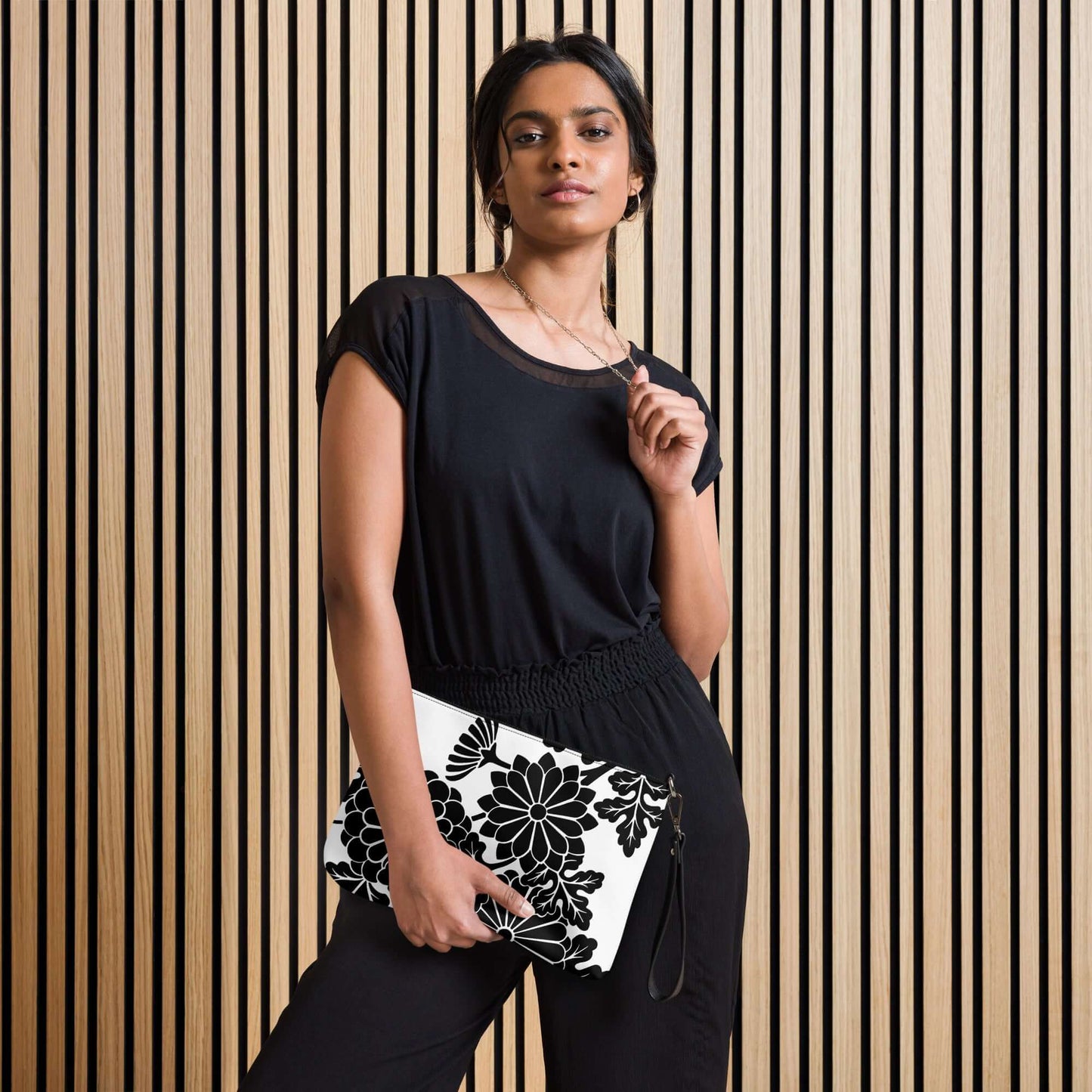 Model holding Umhängetasche N°1 with floral pattern, highlighting versatile black top and pants against wooden backdrop. Fashion and style focus.