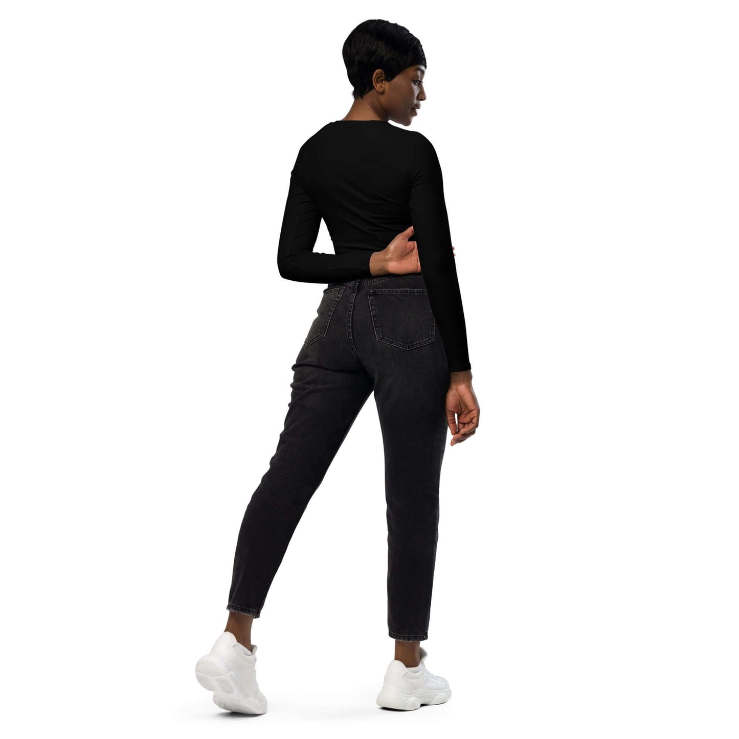 Woman wearing Recyceltes Langarm Crop Top mit Allover-Druck N°3 in black, showing back view, paired with black jeans and white sneakers.