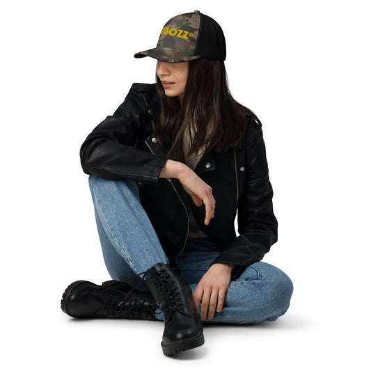 Woman wearing Trucker-Hat im Camouflage-Look N°1, sitting in casual outdoor attire with a black jacket and jeans.