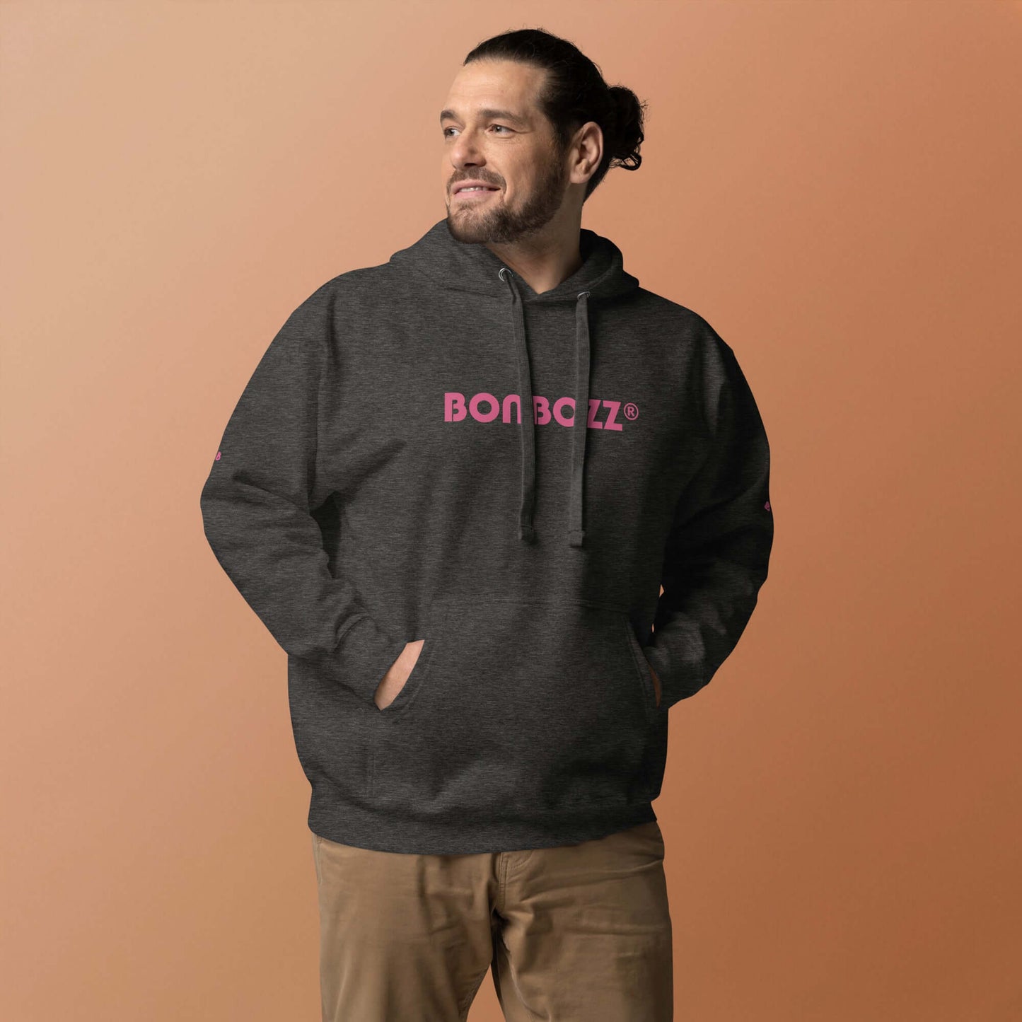 Man wearing a dark grey Unisex-Kapuzenpullover with pink logo, featuring a front pocket and hood, against an orange background.