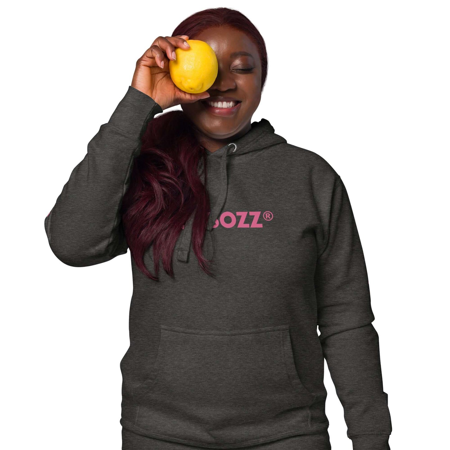 Smiling person wearing a dark grey unisex-Kapuzenpullover with a pink logo, holding a yellow fruit against face.