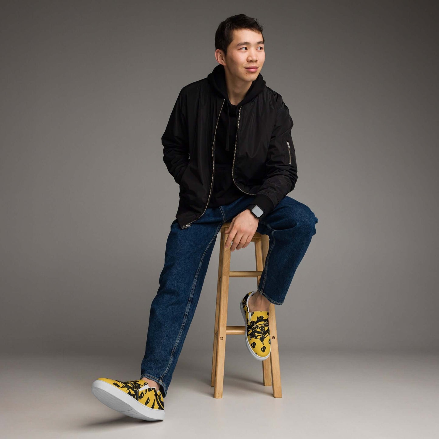 Man wearing Herren-Canvas-Slipper N°1 with casual black jacket and jeans, sitting on a wooden stool, showcasing stylish yellow slip-on shoes.