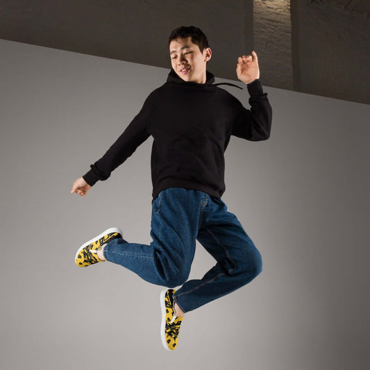 Young man jumping in mid-air wearing yellow floral sneakers, blue jeans, and a black hoodie against a gray background.