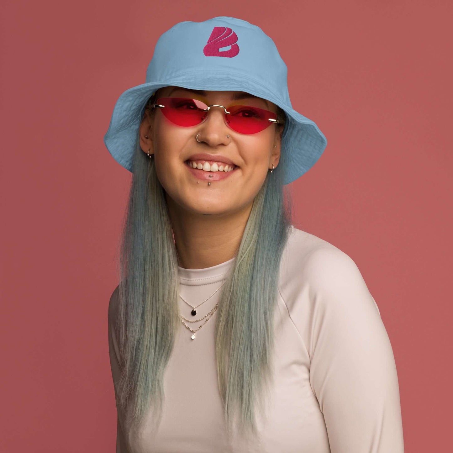 Person wearing Fischerhut aus Bio-Baumwoll-Twill N°1 with bright blue design, smiling in red sunglasses against a pink background.