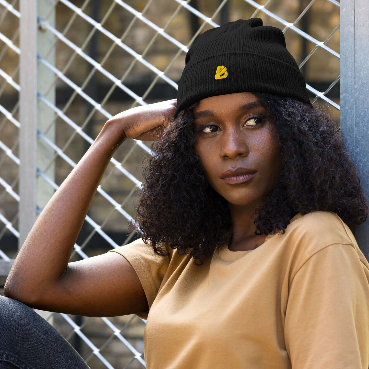 Stylish woman wearing Gerippte Bio-Beanie in black, made from organic cotton, sitting by a metal fence.
