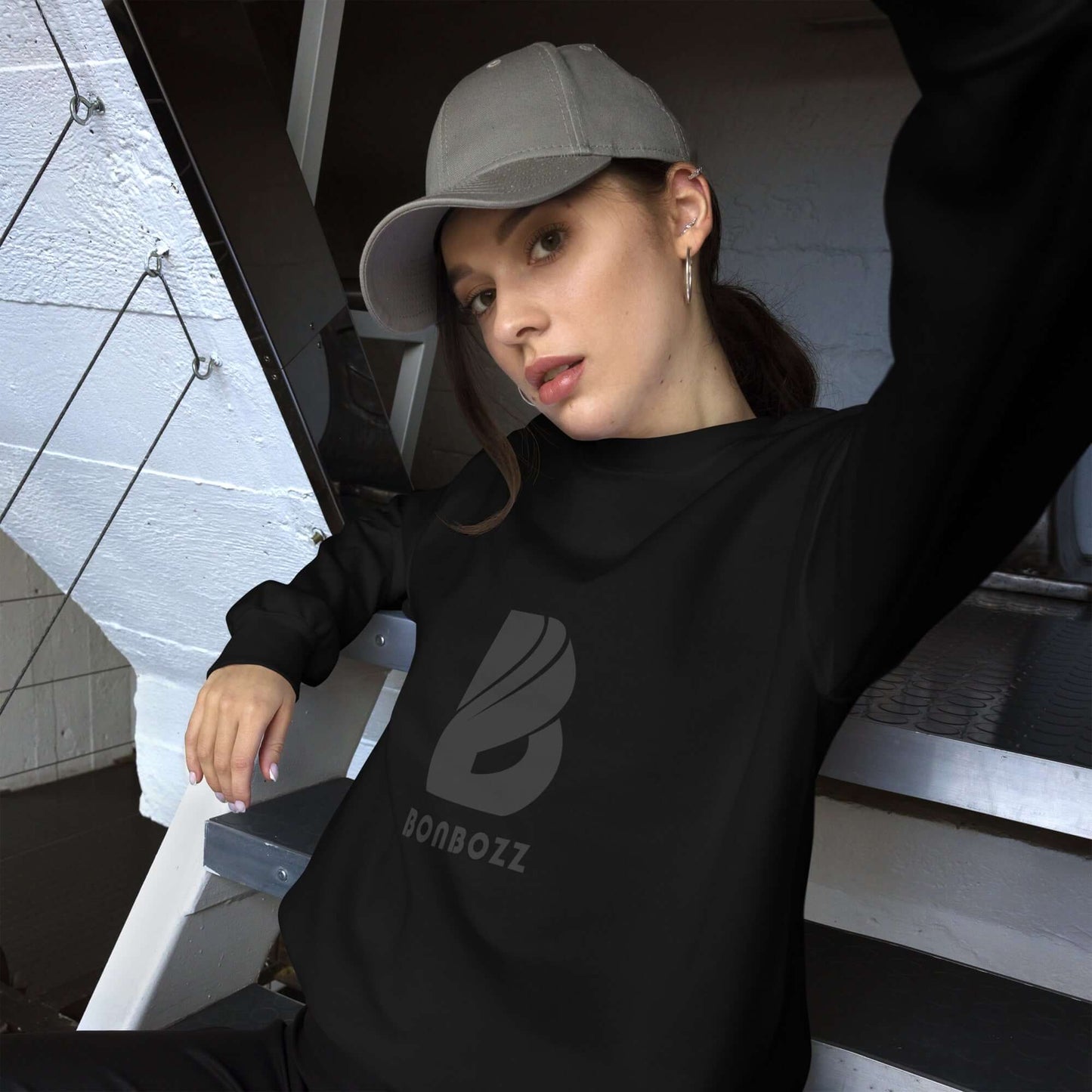 Woman wearing a black Unisex-Pullover and cap, sitting on stairs.