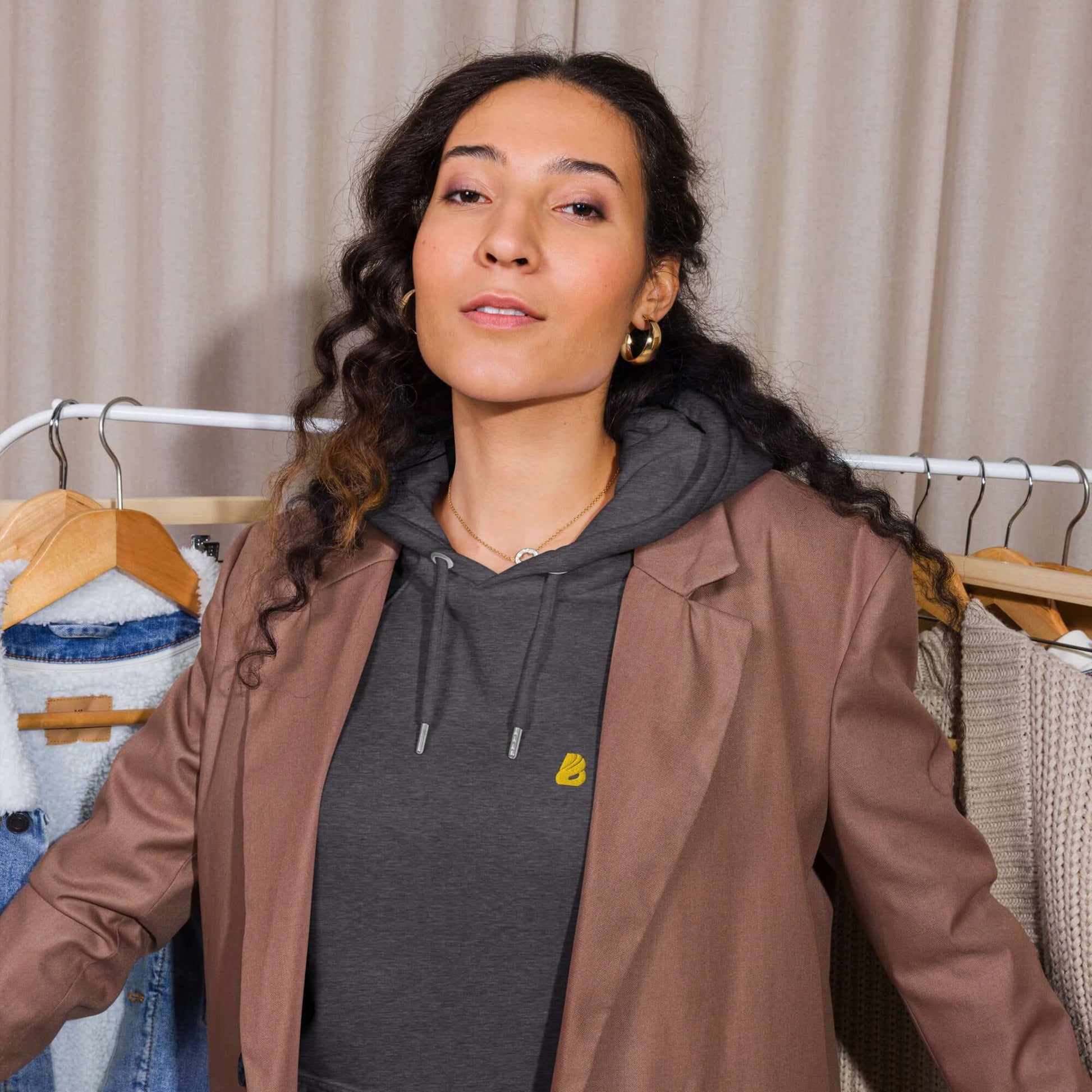 Model wearing a brown blazer over Unisex Bio-Raglan-Hoodie N°5, standing in front of a clothing rack.