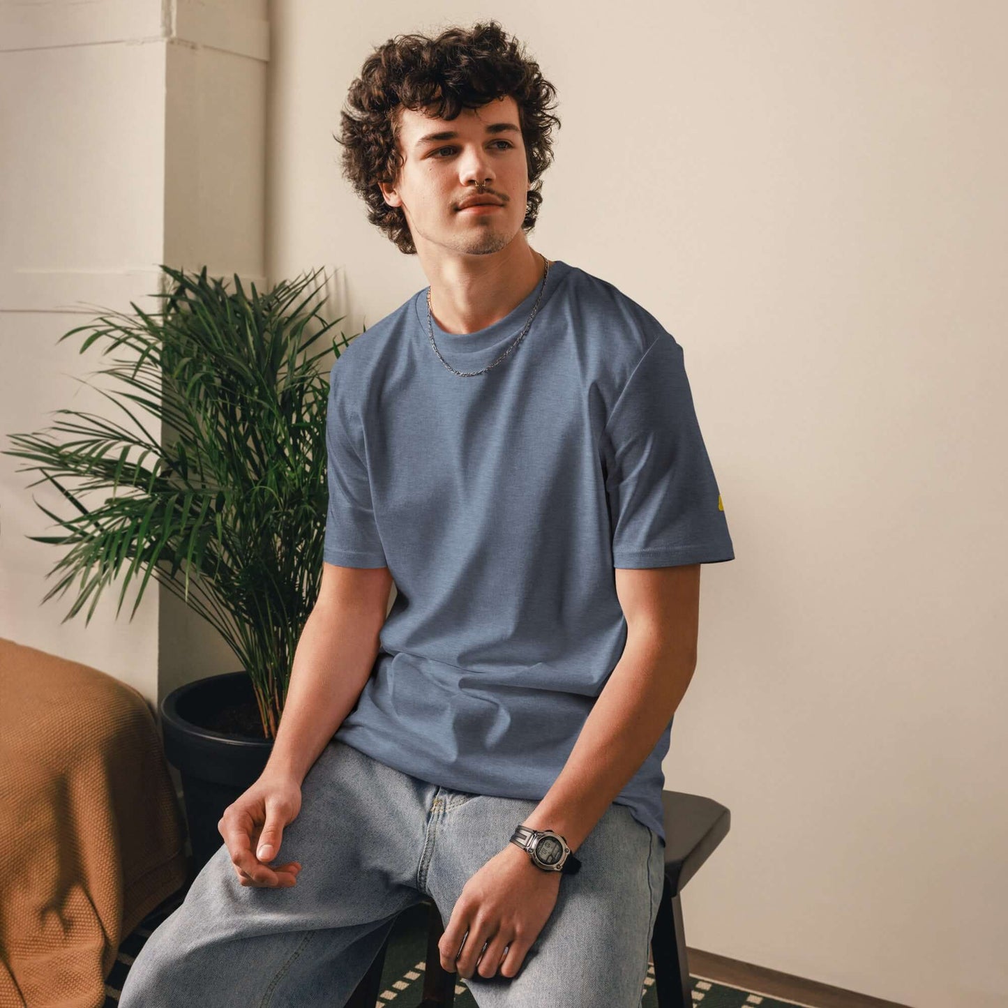 Model wearing a Unisex-Bio-Baumwoll-T-Shirt in blue, sitting indoors next to a plant, showcasing eco-friendly casual fashion.