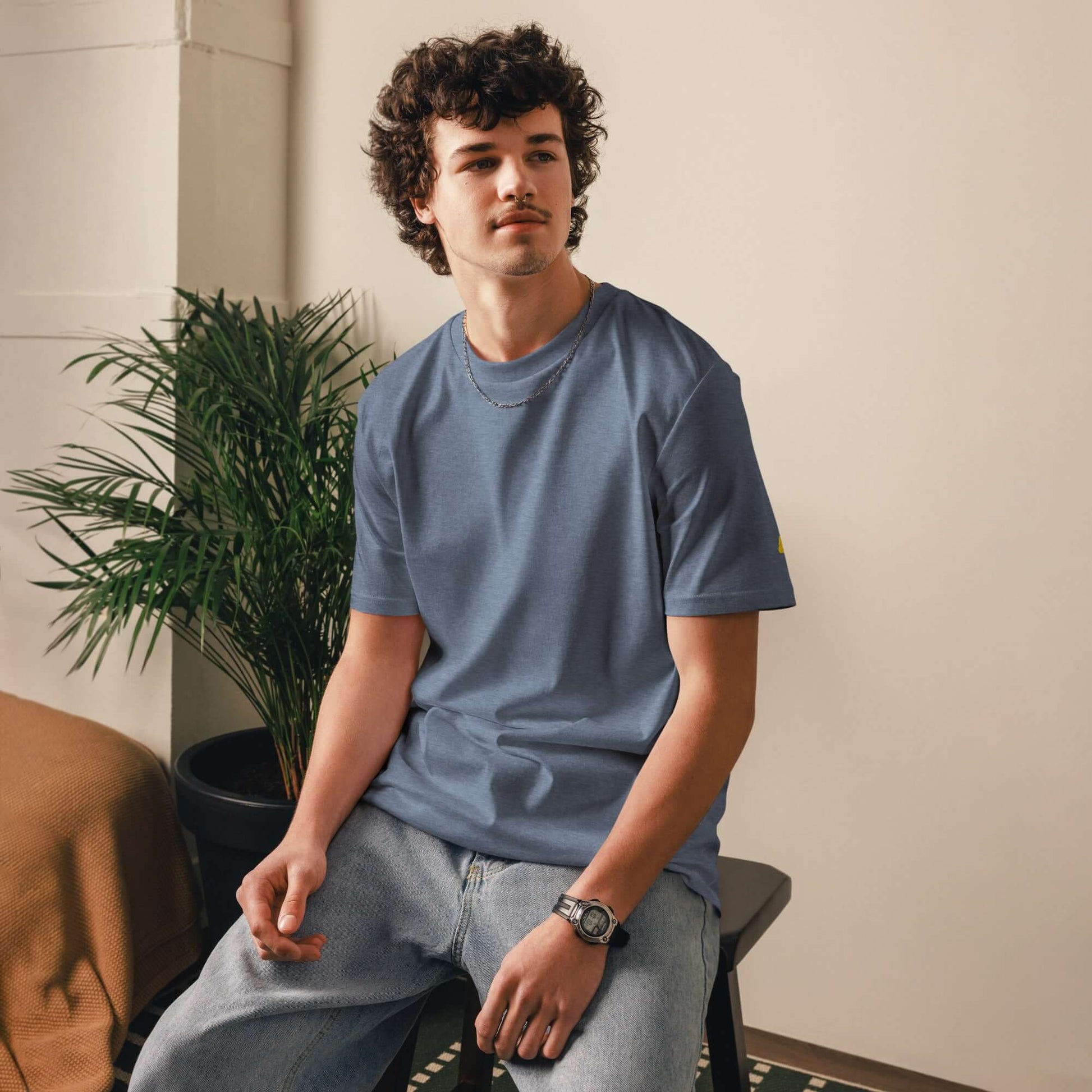 Model wearing a Unisex-Bio-Baumwoll-T-Shirt in blue, sitting indoors next to a plant, showcasing eco-friendly casual fashion.