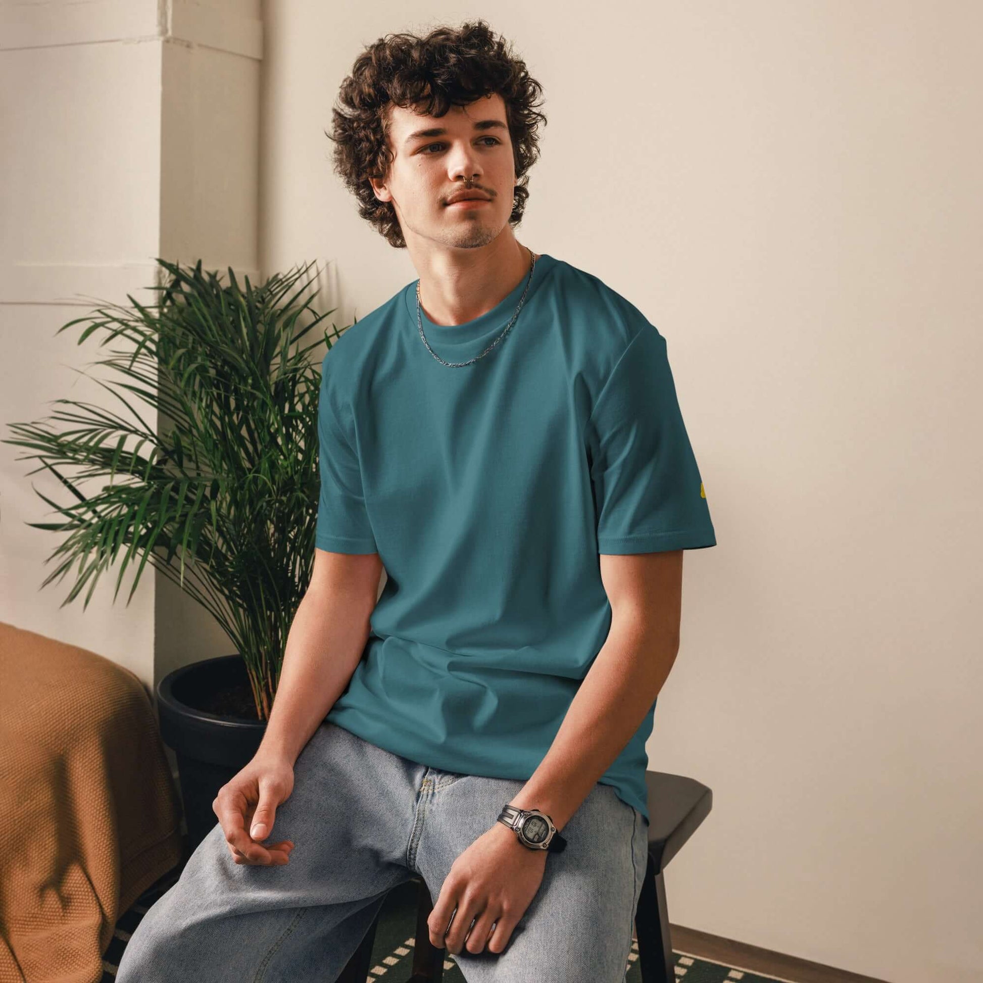 Person wearing a Unisex-Bio-Baumwoll-T-Shirt in teal, sitting indoors beside a potted plant, showcasing eco-friendly fashion.