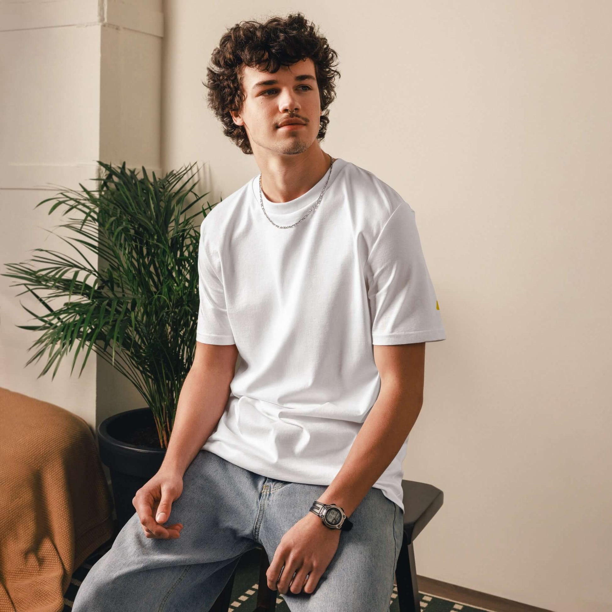 Model wearing a Unisex-Bio-Baumwoll-T-Shirt in a minimalist room setting with a plant backdrop.