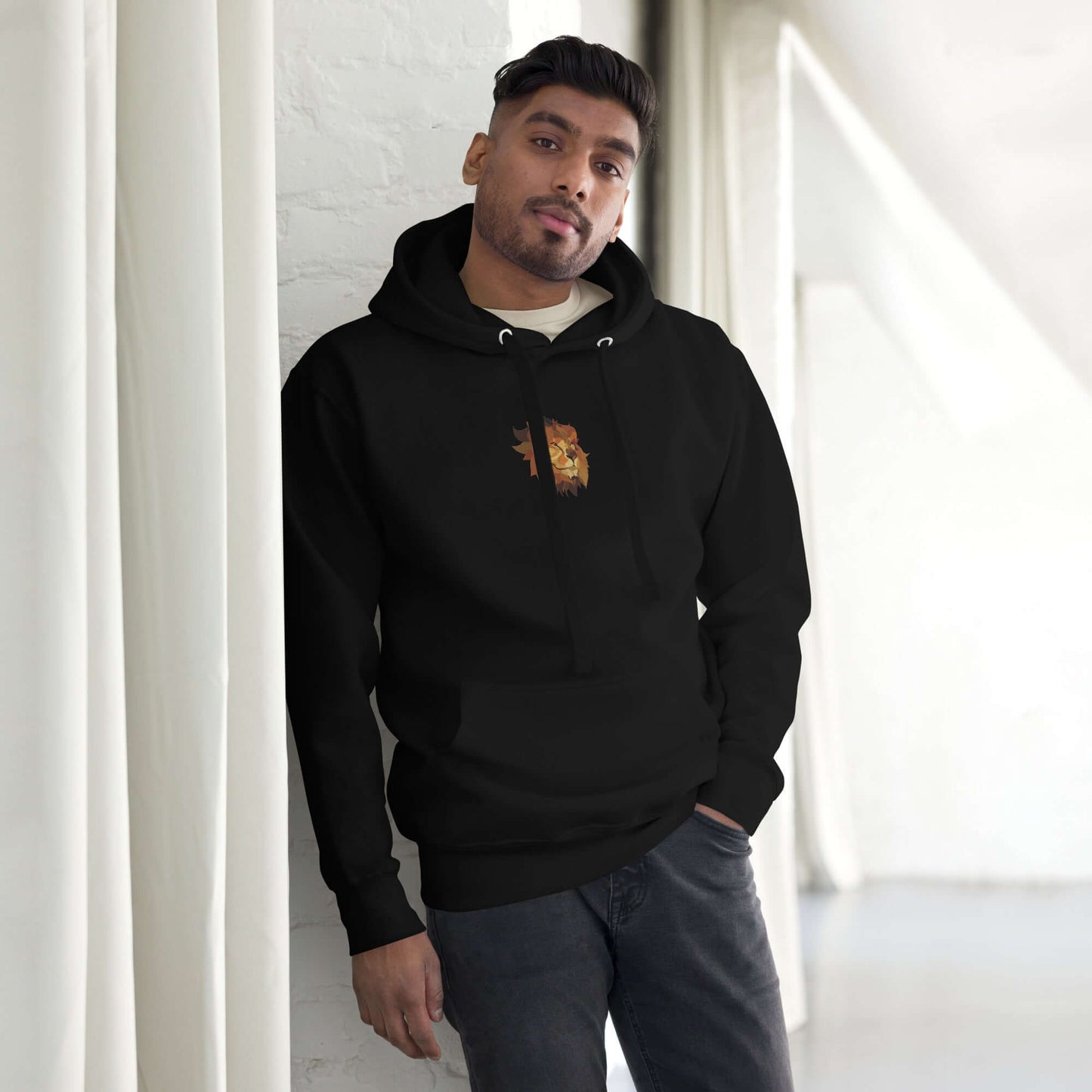 Man wearing Unisex-Kapuzenpullover N°48 in stylish black with kangaroo pocket, leaning against a white wall in a modern room.