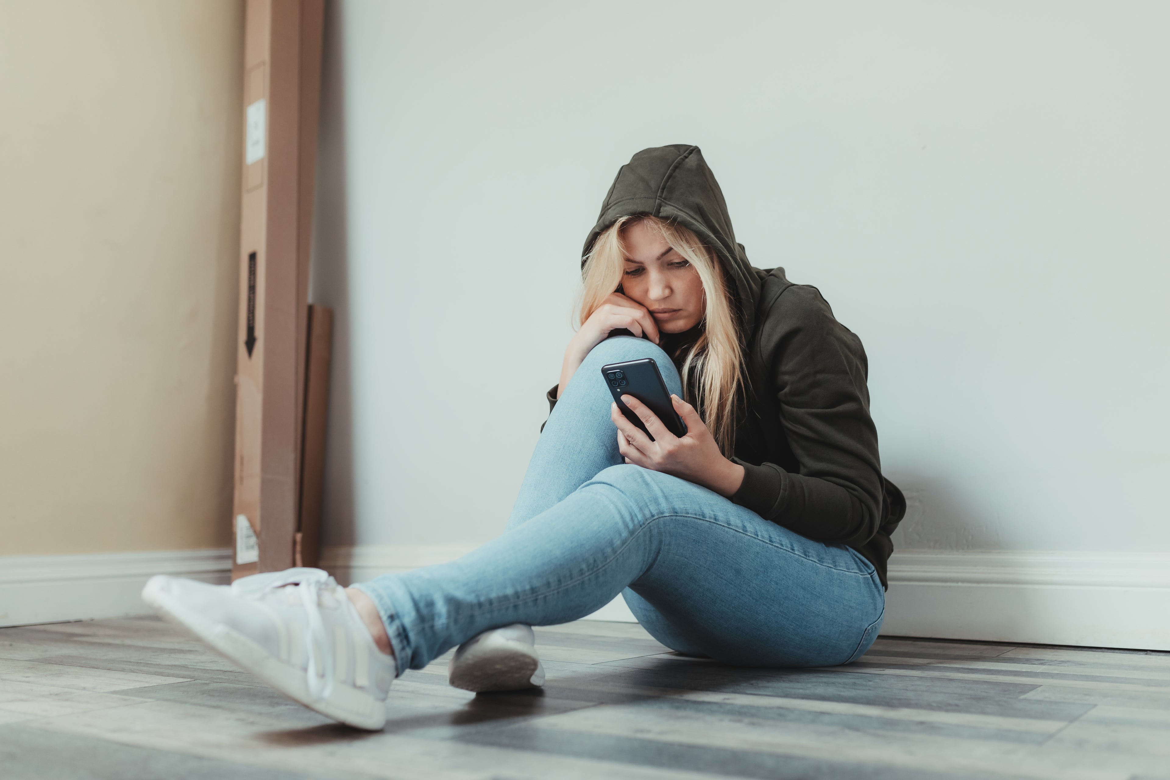 A young woman sitting on the floor in a hoodie, thoughtfully looking at her phone in a minimalist room.