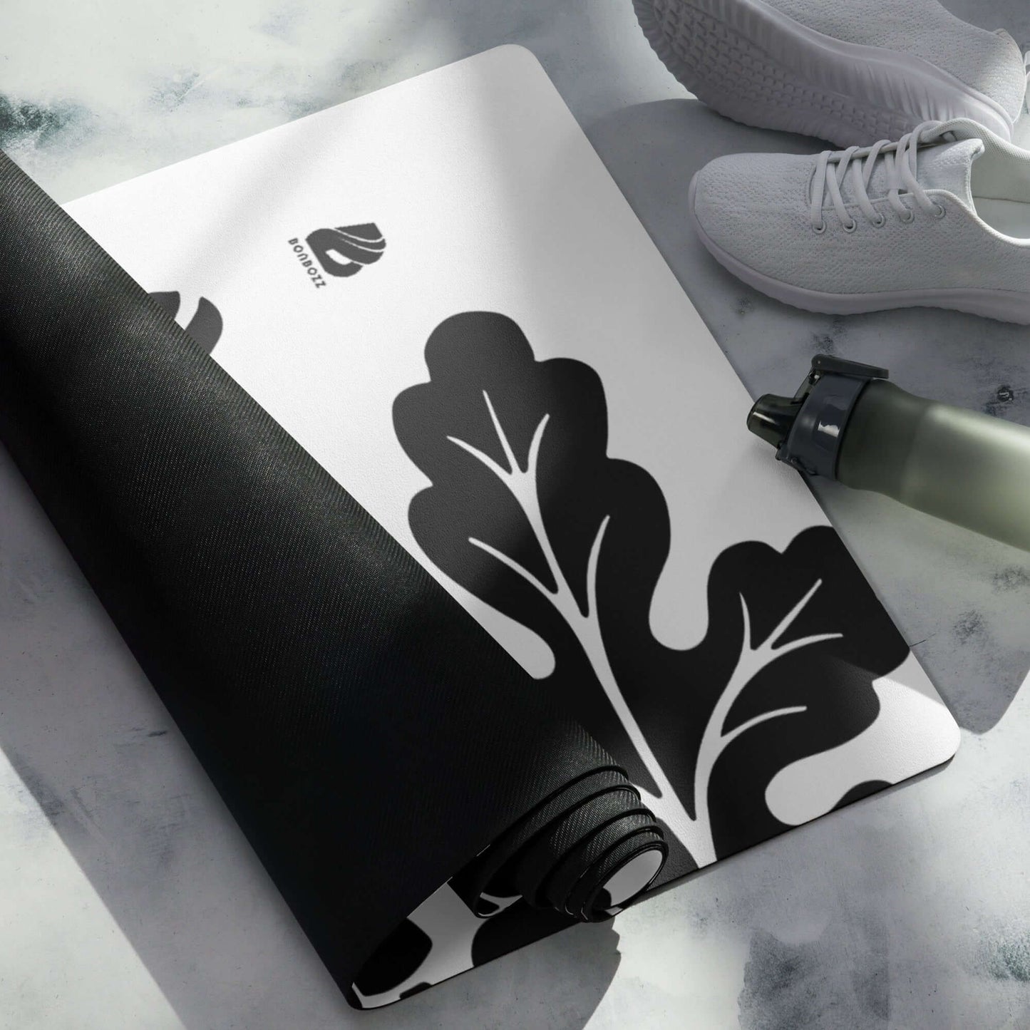 Yoga-Matte N°0 with a black leaf design, rolled on marble surface, shown with water bottle and sneakers. Non-slip, microvelour top.