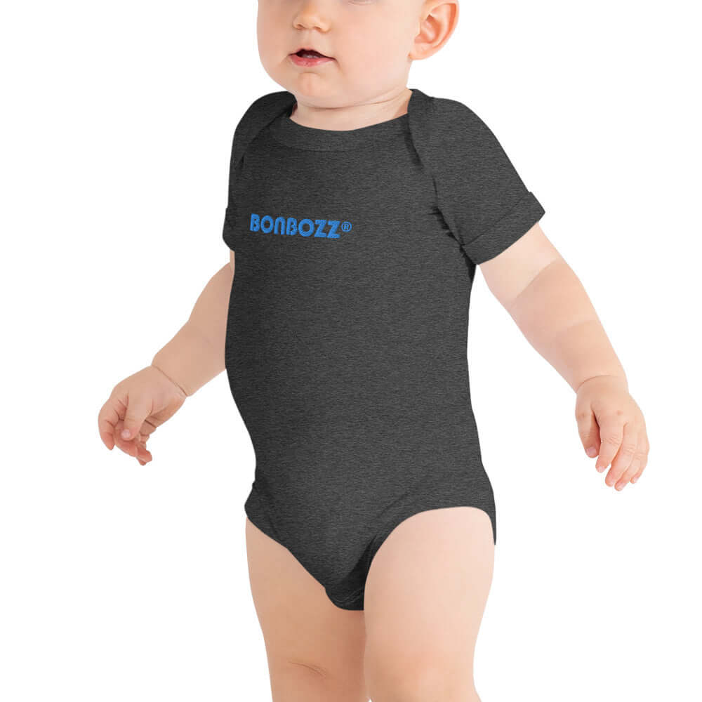 Baby wearing Kurzärmliger Baby-Einteiler N°0 in dark grey with blue text, made from 100% cotton, featuring three snap button closures.