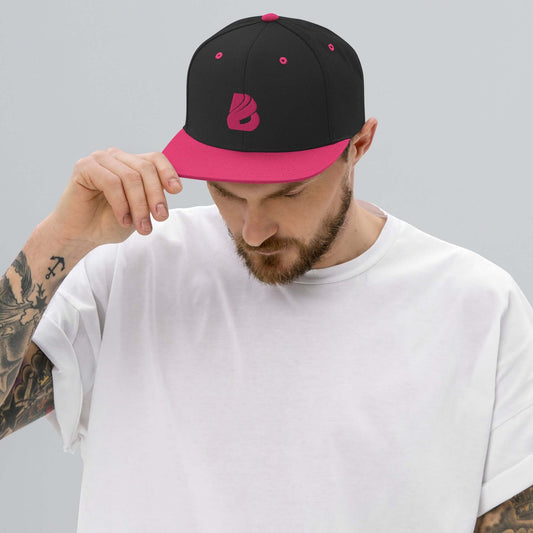 Man wearing Snapback-Cap N°3 with black and pink design, adjusting brim, tattoo visible on arm, gray background.