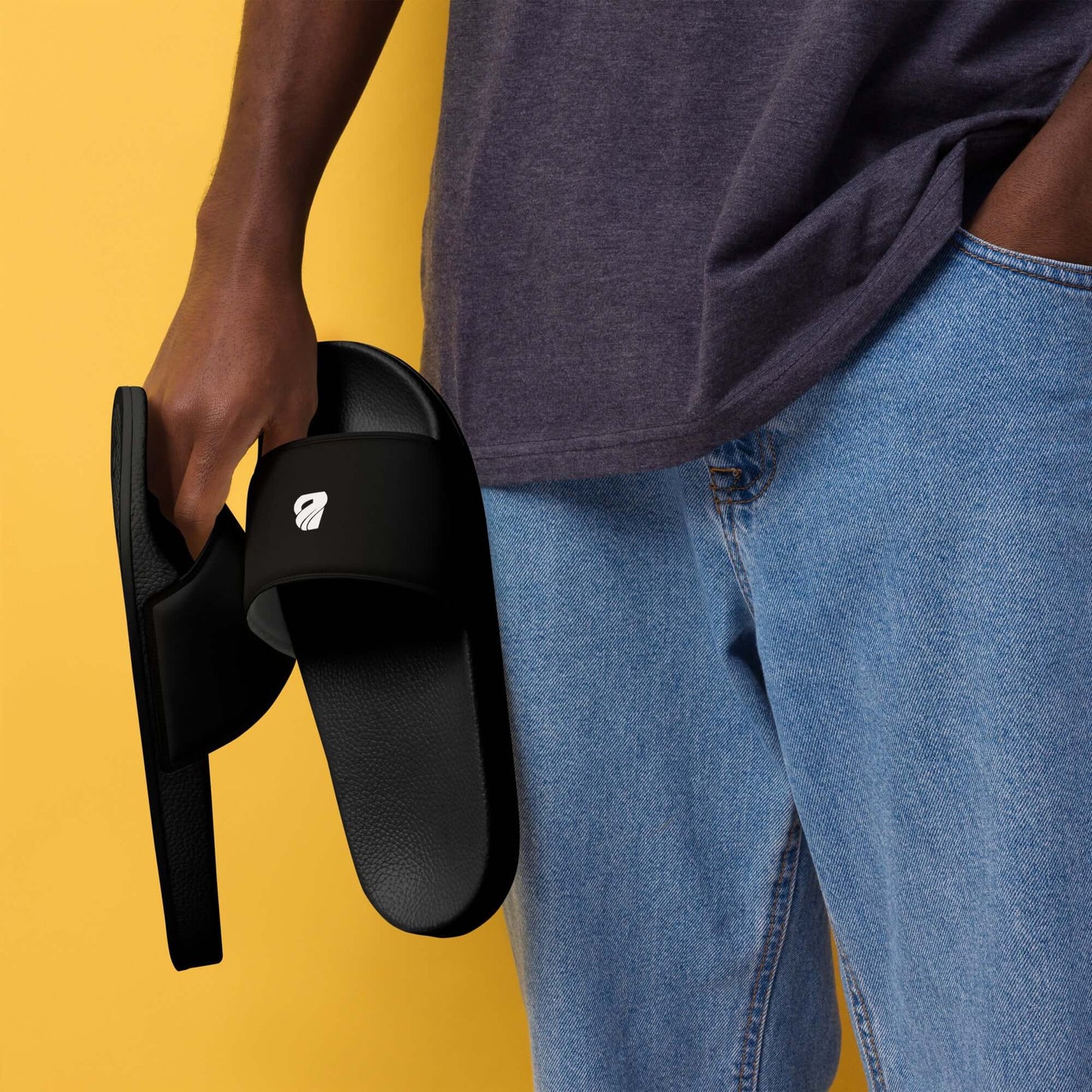 Person holding Herren-Schlappen N°1 sandals, showcasing black padded strap and textured footbed against yellow background and denim jeans.