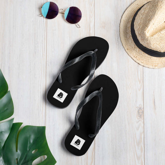 Black Flip-Flops N°8 with printed straps on wooden floor next to sunglasses and hat.