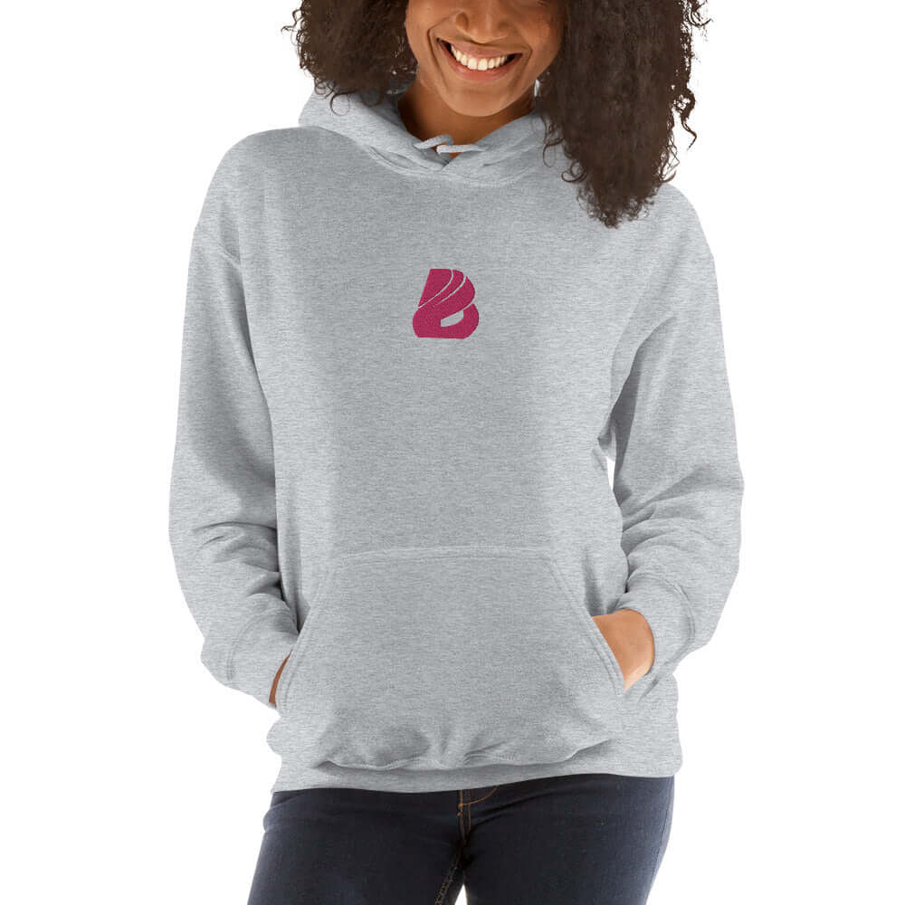 Woman wearing a gray Unisex-Kapuzenpullover N°44 with pink logo, smiling and standing.