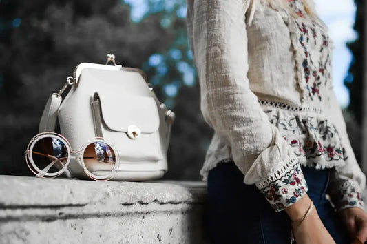 Stylish outfit featuring embroidered blouse, sunglasses, and a white handbag against a natural background.