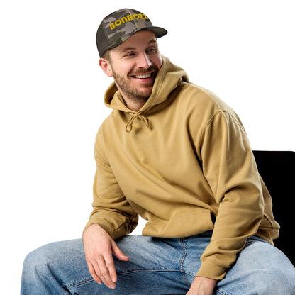 Man wearing Trucker-Hat im Camouflage-Look N°1 with beige hoodie and jeans, smiling and sitting on a chair.