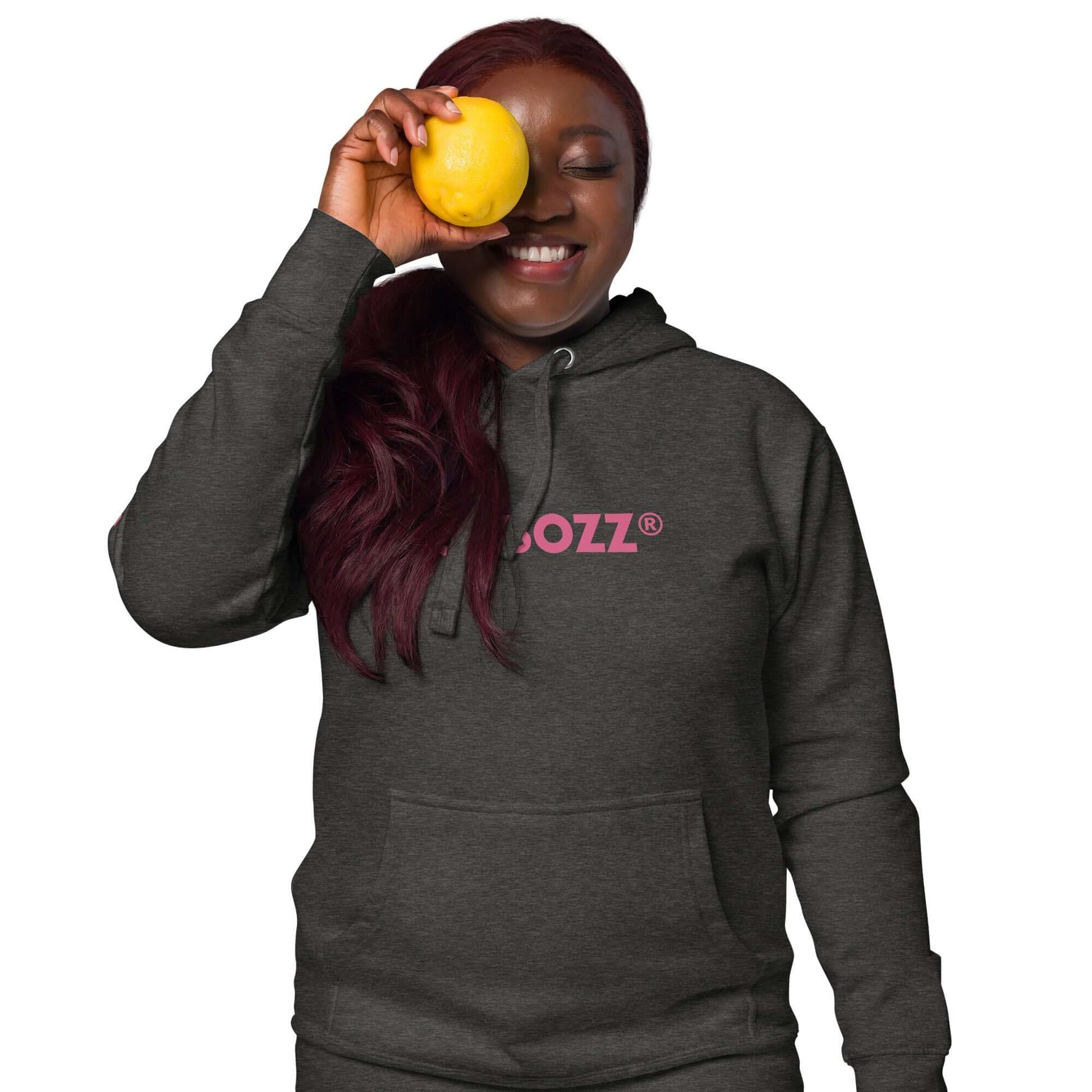 Smiling person wearing a dark grey unisex-Kapuzenpullover with a pink logo, holding a yellow fruit against face.