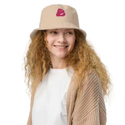 Smiling woman wearing Fischerhut aus Bio-Baumwoll-Twill N°0, showcasing eco-friendly and stylish design in beige with pink logo.