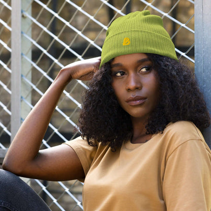 Stylish woman wearing a green Gerippte Bio-Beanie against a metal fence backdrop.