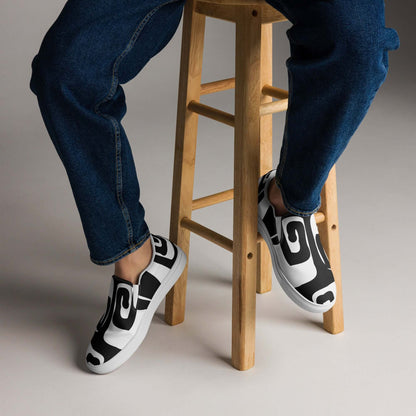 Herren-Canvas-Slipper N°0 worn by a person sitting on a wooden stool, showcasing stylish design and comfortable fit with patterned black and white canvas.