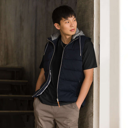 Young man wearing a stylish black vest over a hoodie, standing indoors and leaning against a wall with a thoughtful expression.