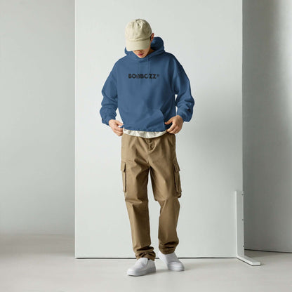 Person wearing Unisex-Kapuzenpullover N°46 in blue with beige cargo pants and cap, standing in a minimalist room.