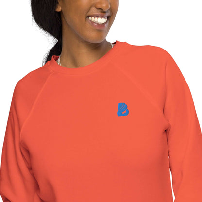 Person wearing Unisex Bio-Raglan-Pullover N°1 in vibrant orange with blue logo, showcasing style and comfort in organic cotton.